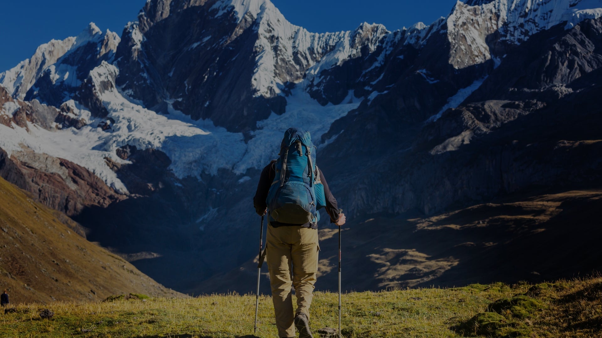 Hiker with a backpack and walking sticks standing in front of snow-capped mountains.